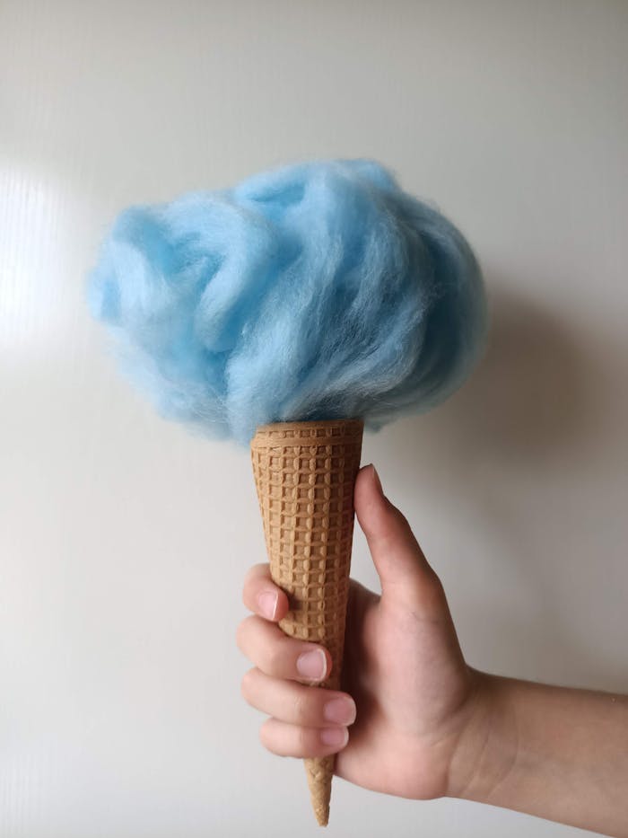 Hand holding fluffy blue cotton candy in a waffle cone against a white background.