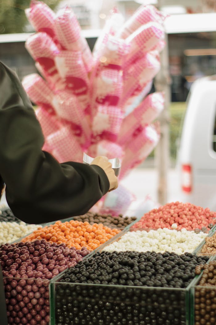 Person selecting colorful candies at an outdoor market stall with cotton candy in the background.